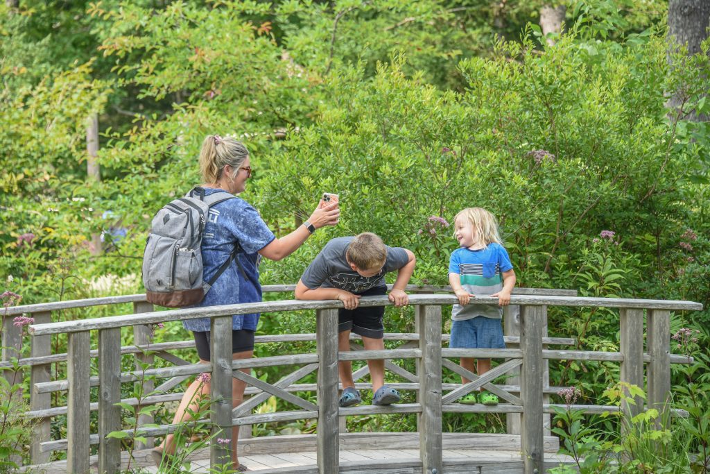 Two smiling children stand on a wood bridge, set in a garden. A parent shows the children a photo on their phone.